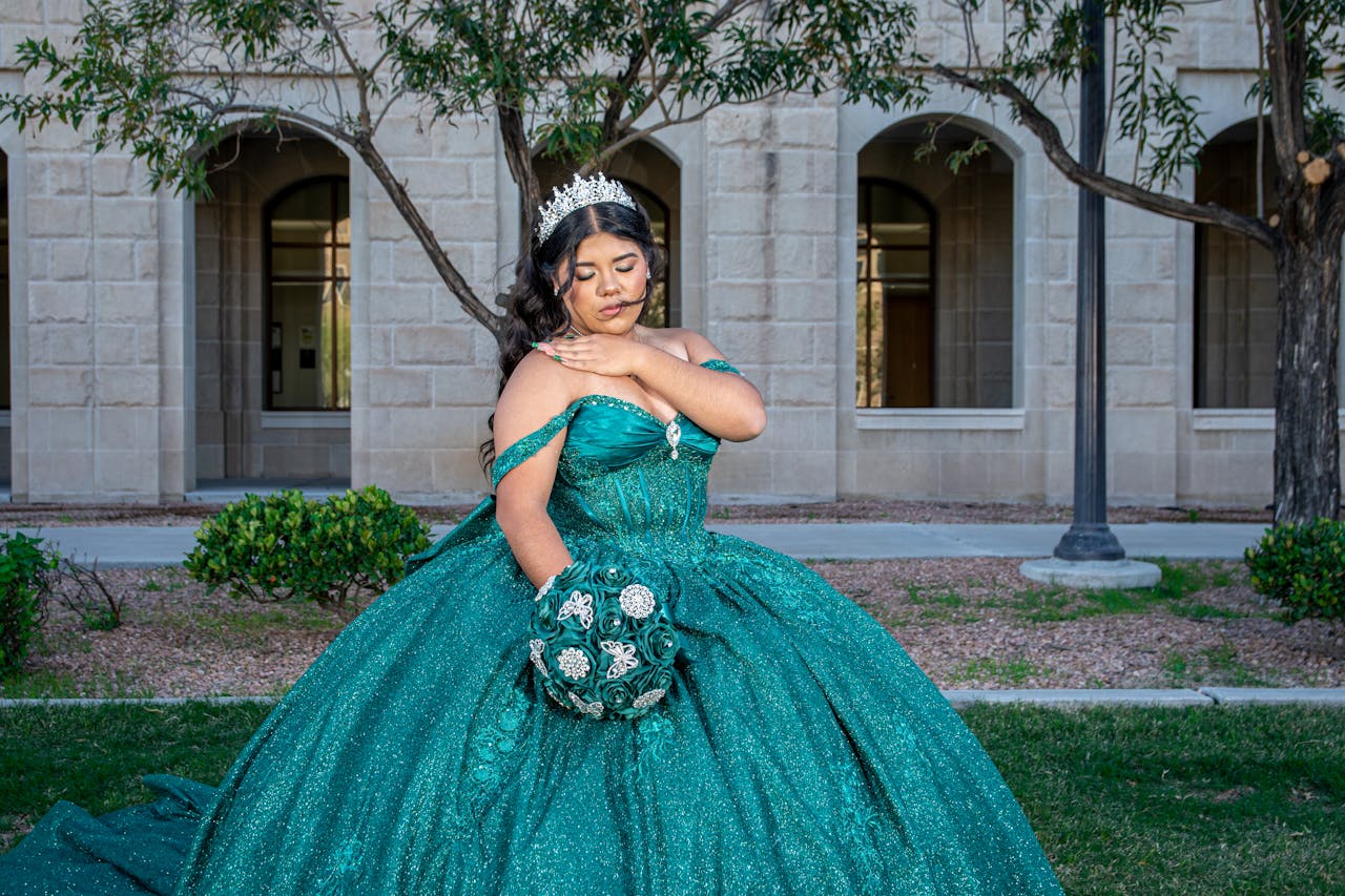 Elegant young woman in a teal quinceañera dress with a tiara poses outdoors.